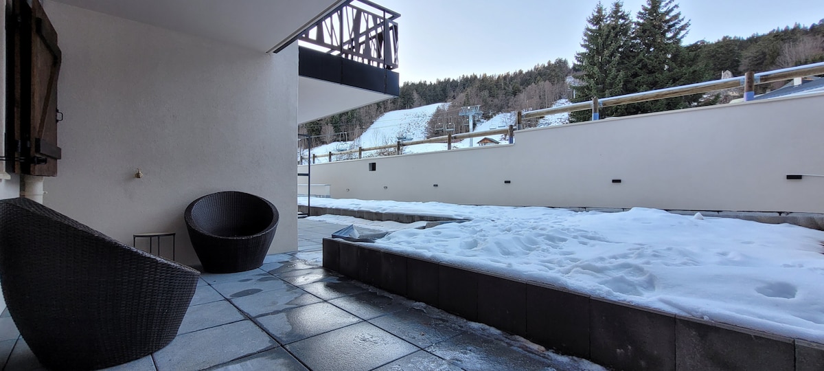 An outdoor terrace with stone flooring is visible, complemented by two wicker chairs. Snow covers the ground, and the surrounding landscape features trees and a ski slope in the distance. A minimalist design enhances the space's modern appeal.