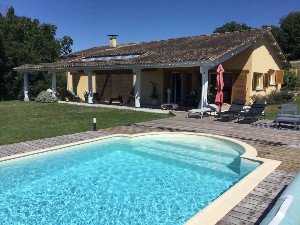 The villa features a private pool surrounded by a spacious wooden deck, complemented by sun loungers and a large umbrella. The exterior showcases a single-story structure with wooden shutters, a sloped roof, and a manicured lawn, all set against a backdrop of greenery.