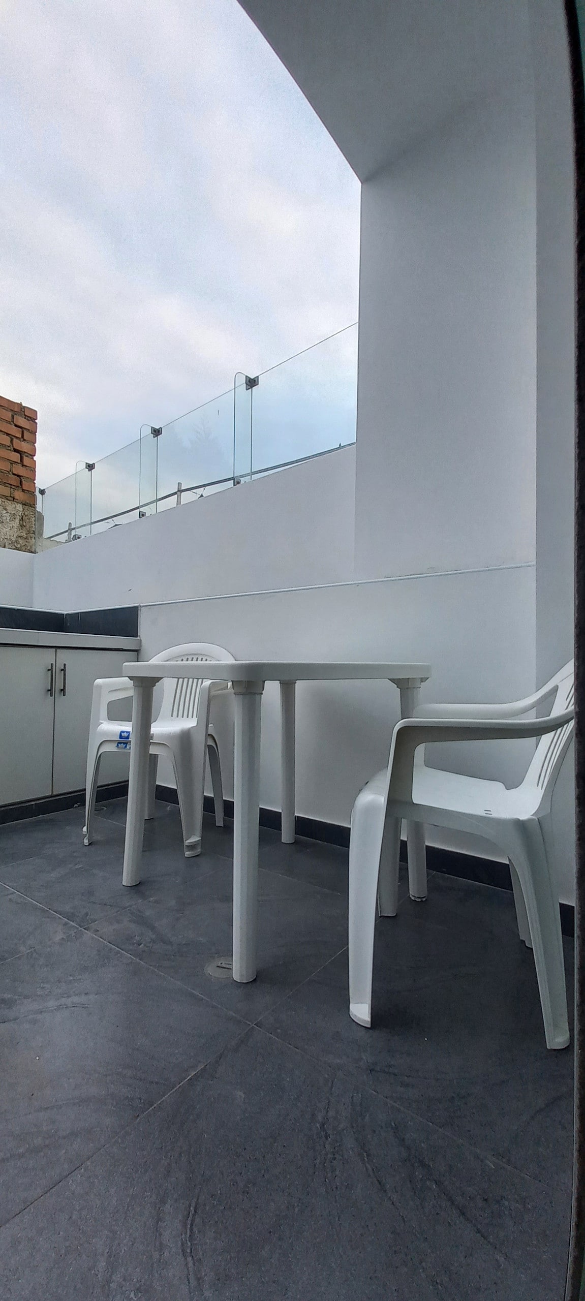 A small outdoor terrace is visible, featuring a table and two white plastic chairs. The space is enclosed by a glass railing, allowing light to flow in while providing an unobstructed view. The floor is tiled in dark grey, contributing to a modern aesthetic.