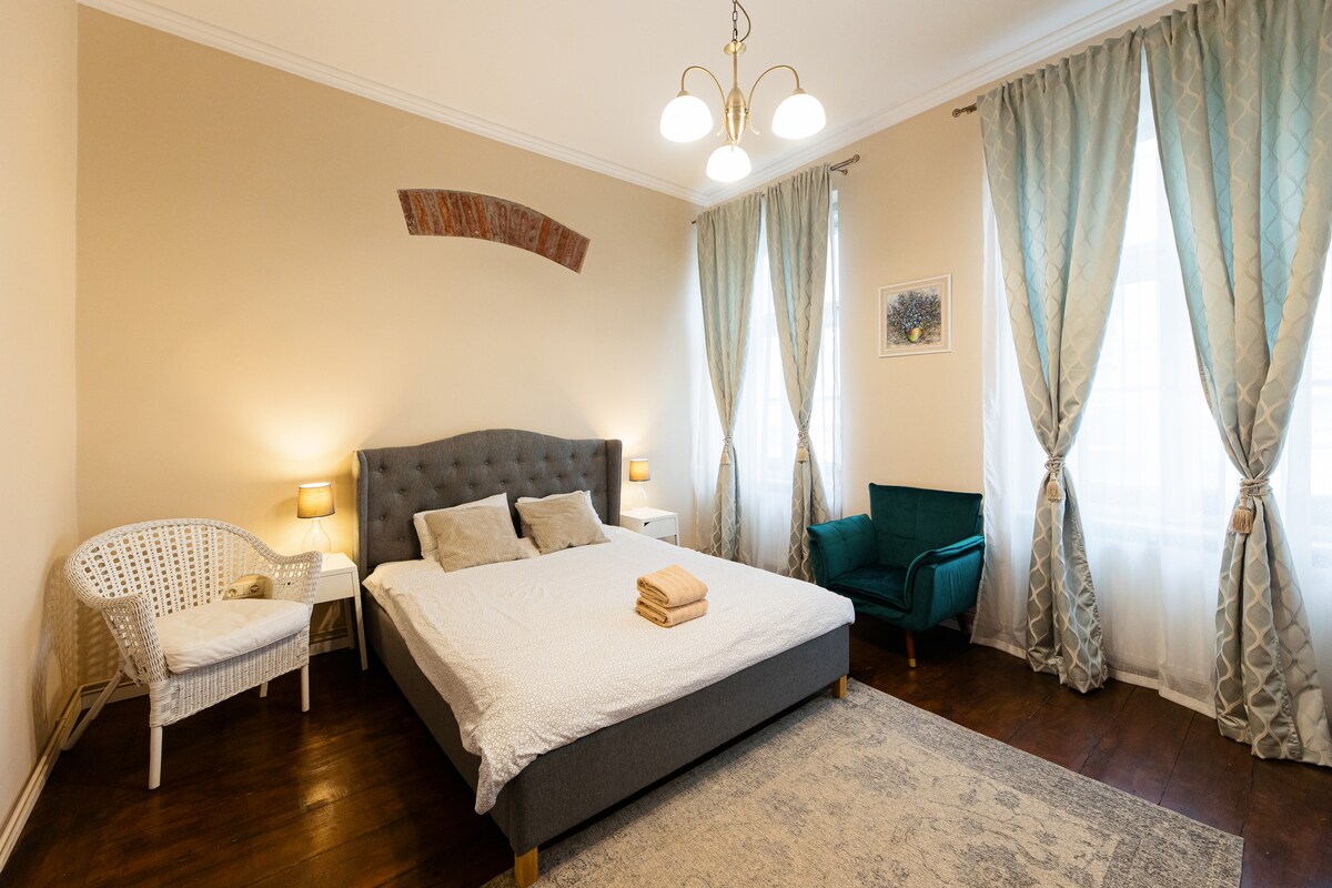 A spacious bedroom is presented with a large bed featuring soft bedding. Two bedside lamps provide gentle illumination. A cozy armchair is positioned near a window draped with sheer curtains, while a decorative rug enhances the warm wooden flooring.