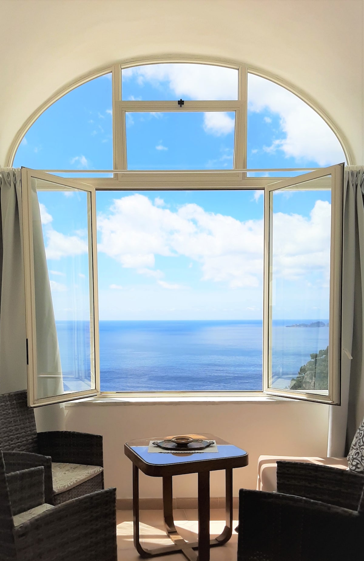 A large arched window opens to reveal a breathtaking sea view beneath a clear blue sky. Two wicker chairs are positioned near a small table, inviting relaxation while enjoying the scenery. Soft curtains frame the window, enhancing the view of the tranquil waters.