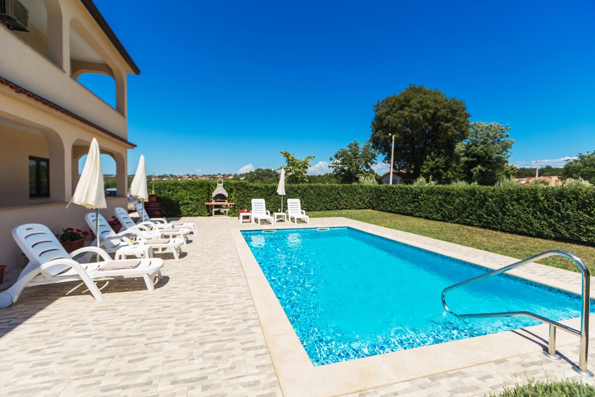 A private pool surrounded by a well-maintained garden features comfortable lounge chairs and outdoor umbrellas. The pool area is situated under clear blue skies, offering a serene environment for relaxation.