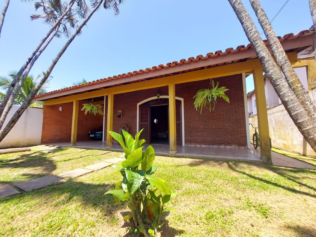 A single-story house is presented, featuring a red brick façade and a tiled roof. The front yard showcases a neatly maintained grassy area with a small green plant in view. Palm trees create a natural border, while a bicycle is positioned near the entrance.