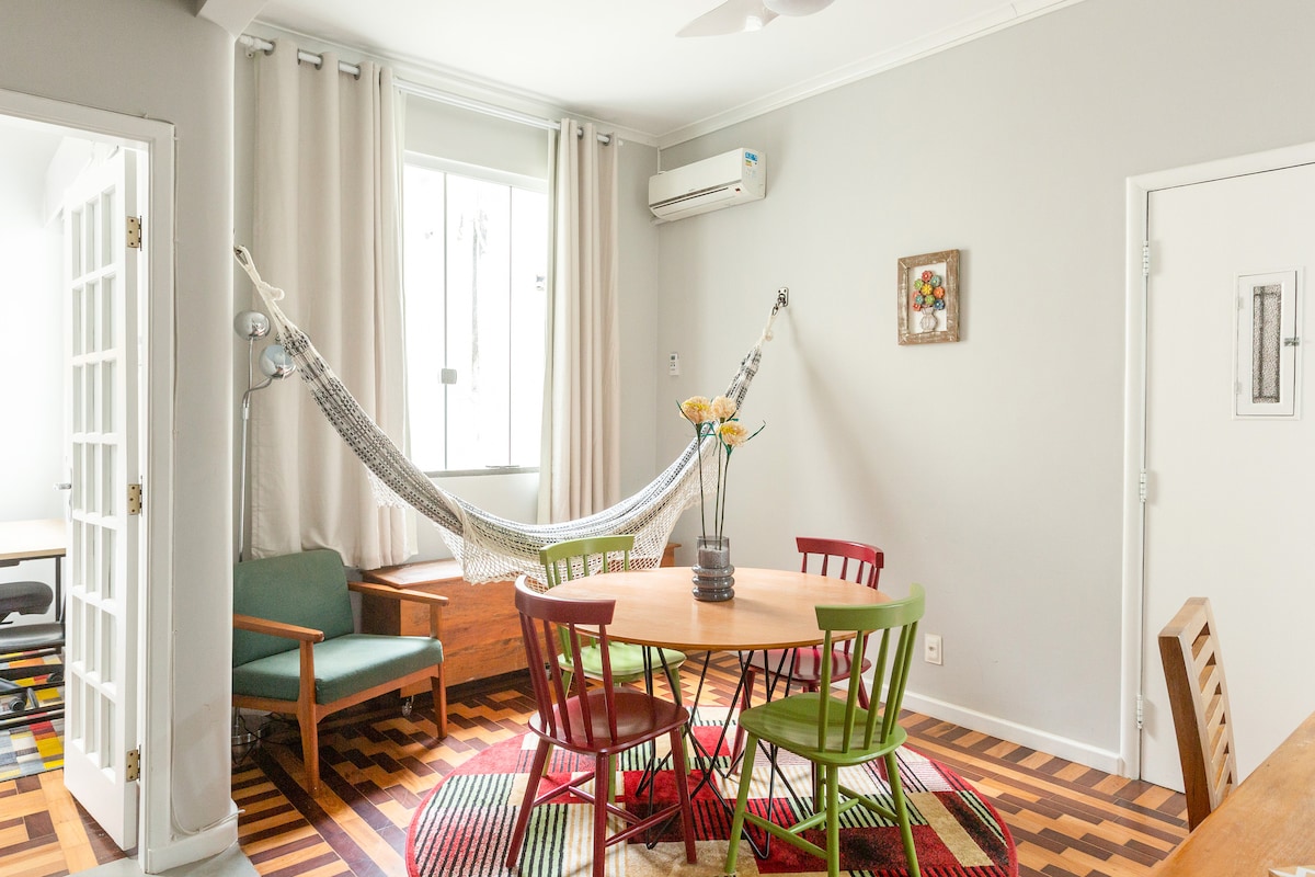 A dining area features a round table surrounded by colorful chairs in red, green, and brown. Light hardwood flooring complements the room, which includes a hammock hung in a corner. A vase with flowers adorns the table, and a window is partially covered by light curtains.