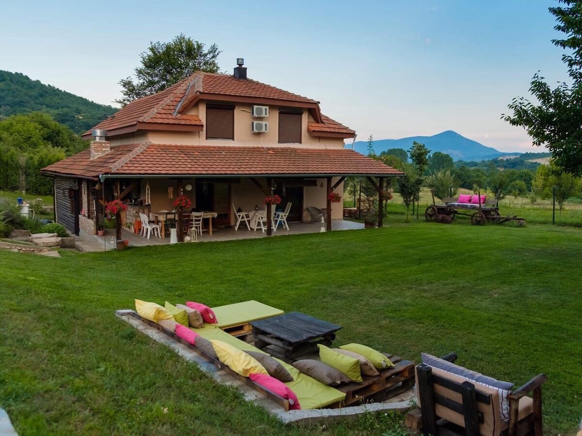 The exterior of the house features a spacious porch adorned with blooming flowers and outdoor seating arrangements. A well-maintained lawn stretches out in front, complemented by lounge cushions arranged on the grass. Rolling hills and trees provide a serene backdrop.