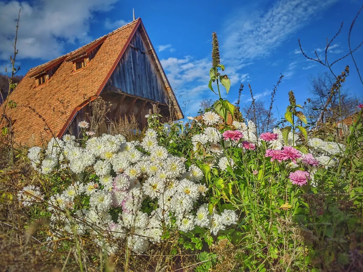 A cluster of vibrant flowers in shades of white and pink occupies the foreground, set against a rustic wooden building with a steep, red-tiled roof. The blue sky above is dotted with wispy clouds, enhancing the peaceful rural setting.