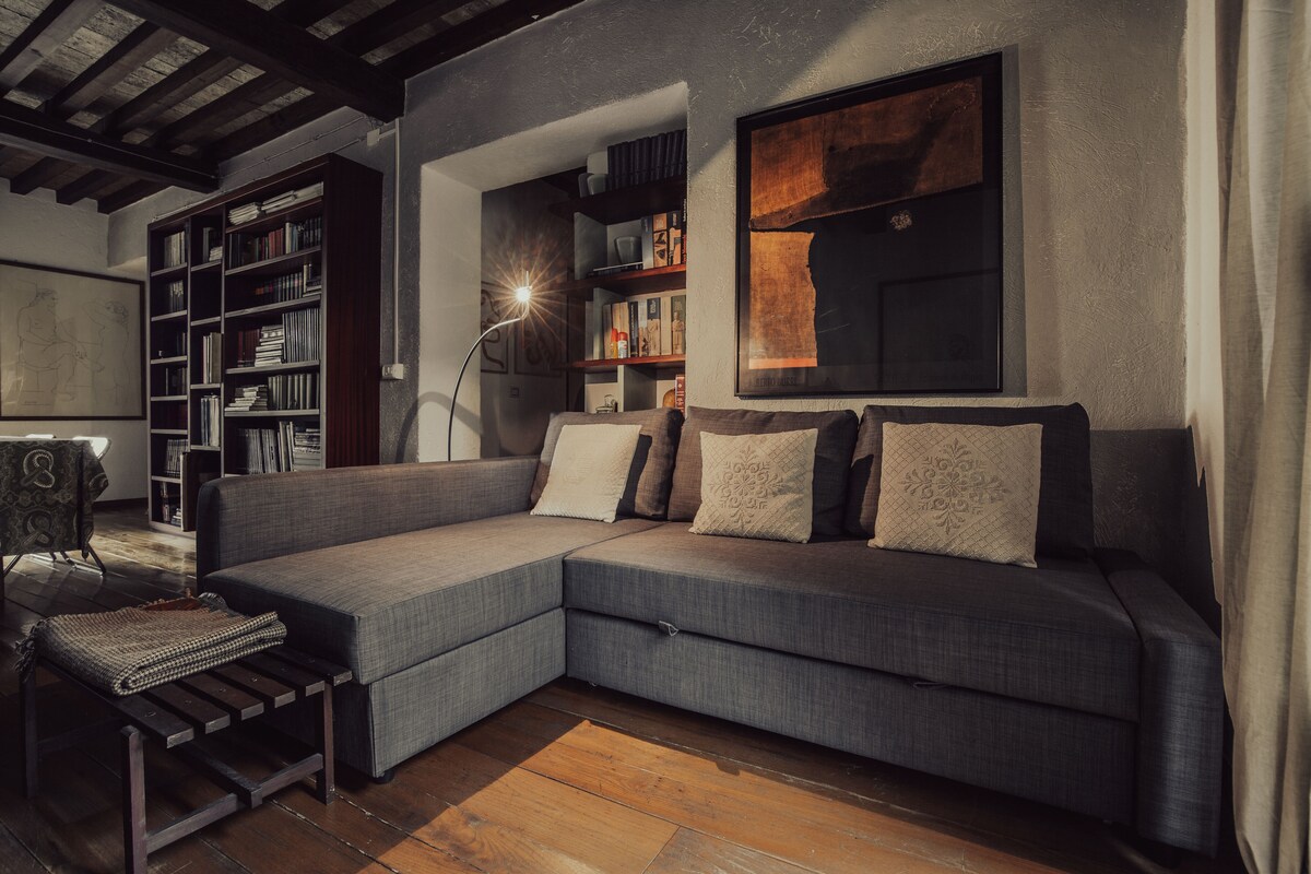 A cozy seating area features a modern gray sectional sofa adorned with decorative pillows. A minimalist floor lamp provides soft lighting beside the sofa. In the background, bookshelves are filled with books, creating a welcoming atmosphere for relaxation.