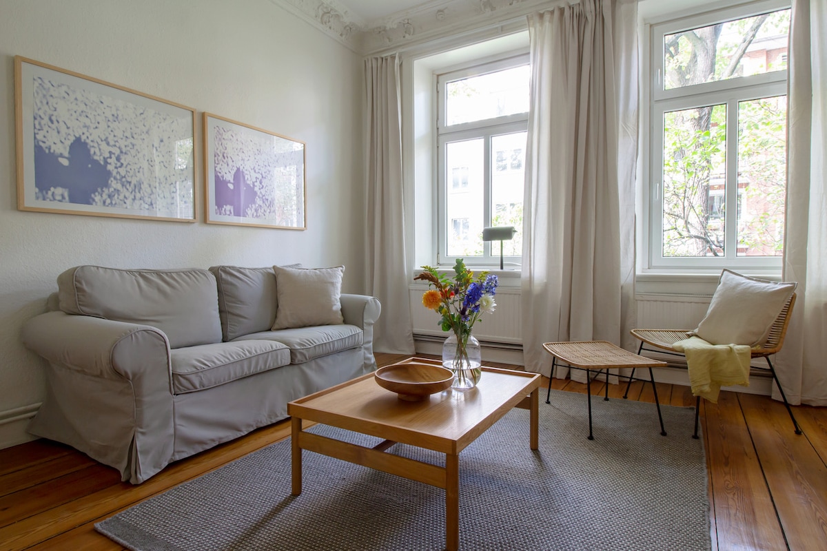 A brightly lit living area features a light-colored sofa beside a stylish chair. A wooden coffee table occupies the center, while a decorative bowl holds colorful flowers. Soft curtains frame the windows, allowing natural light to create a welcoming environment.