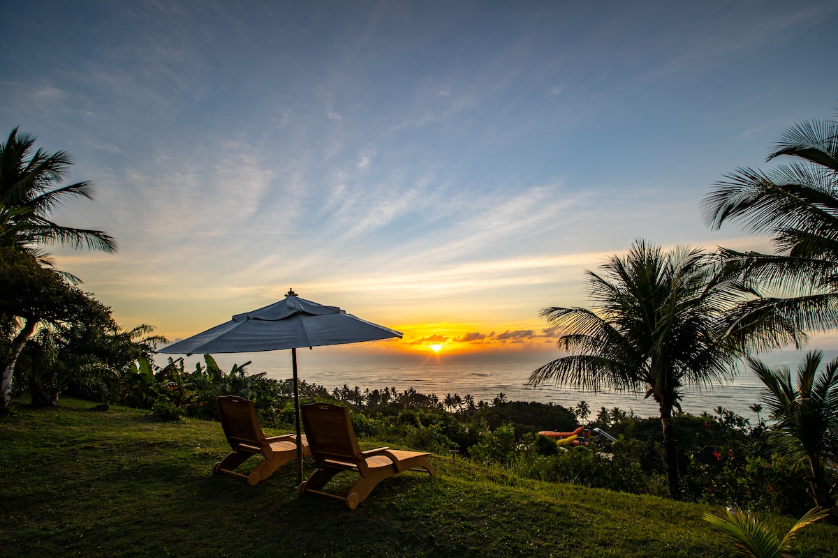 Two lounge chairs are placed under an umbrella on a grassy area, overlooking a panoramic view of the coastline. The horizon features a vibrant sunrise, while palm trees frame the scene, enhancing the natural setting.