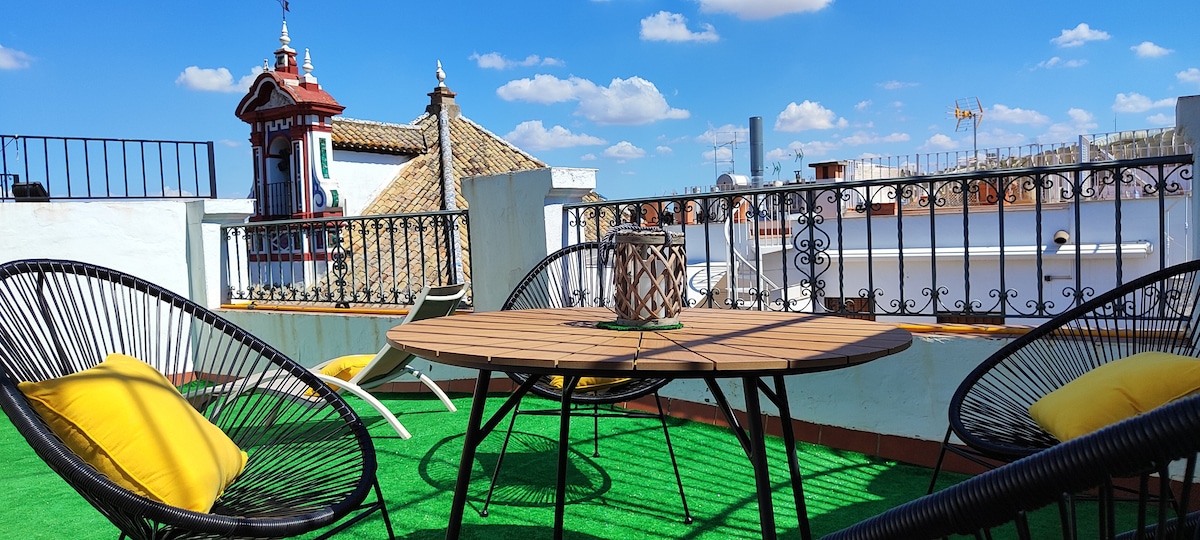 A private terrace is presented with a table surrounded by four chairs, complemented by bright yellow cushions. The space features green grass-like flooring and offers views of nearby rooftops and architectural details under a blue sky with scattered clouds.