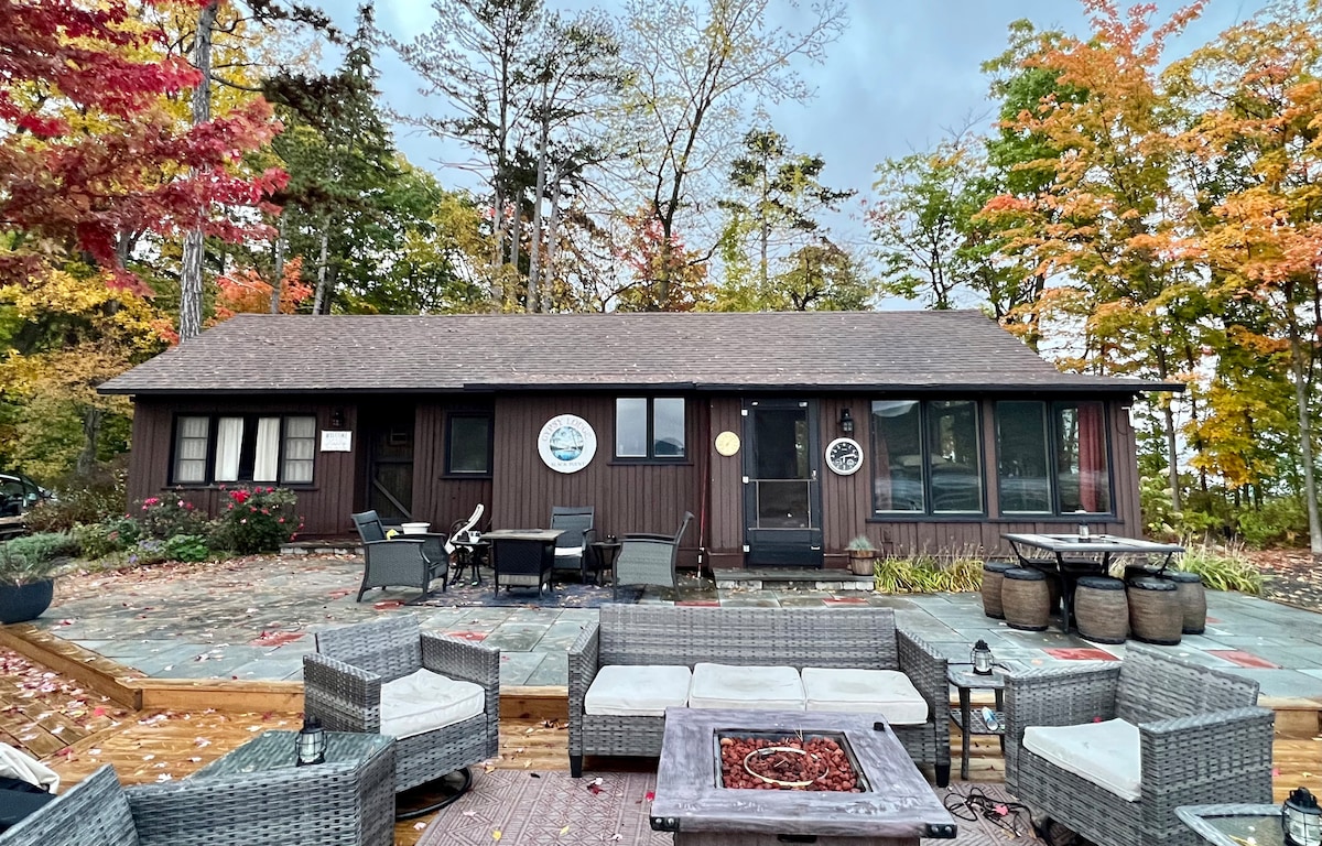 A charming lodge exterior is framed by vibrant autumn foliage, presenting a cozy seating area with wicker chairs and a fire pit. Stone and wooden decks provide space for relaxation, while large windows offer views of the surrounding nature.