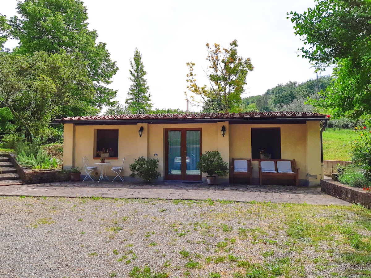 The cottage is positioned amidst a lush garden, featuring a stone pathway leading to the entrance. A rustic facade is complemented by wooden benches and white outdoor furniture, surrounded by trees and greenery that provide a serene rural backdrop.
