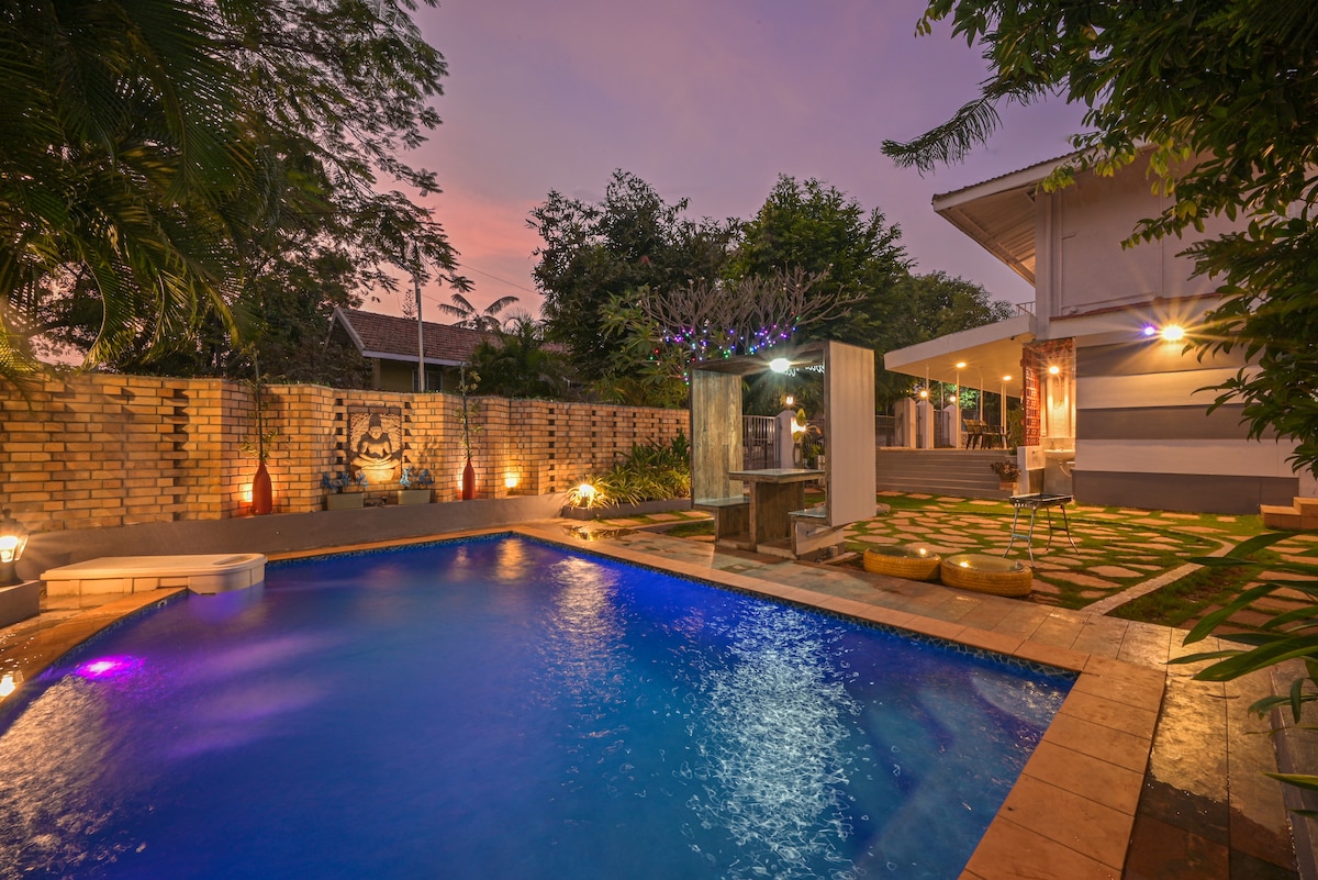 An outdoor space features a private pool illuminated by soft lighting, surrounded by landscaped greenery. A barbecue area and seating arrangements are visible, with a serene garden backdrop. The warm evening sky adds to the tranquil ambiance, making it suitable for relaxation and gatherings.