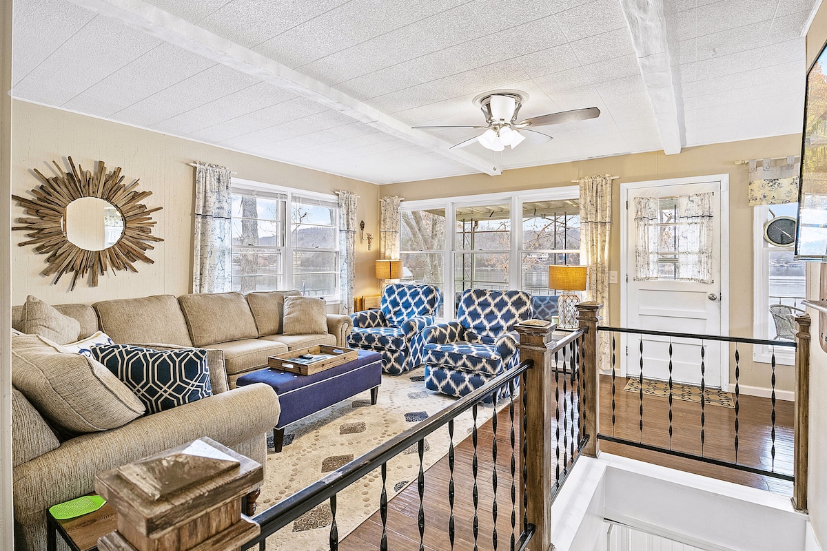 A welcoming living area features a large sectional sofa and two accent chairs with geometric patterns. Natural light filters through multiple windows, highlighting the warm tones of the furnishings. A coffee table is centered on a light area rug, complementing the cozy atmosphere.