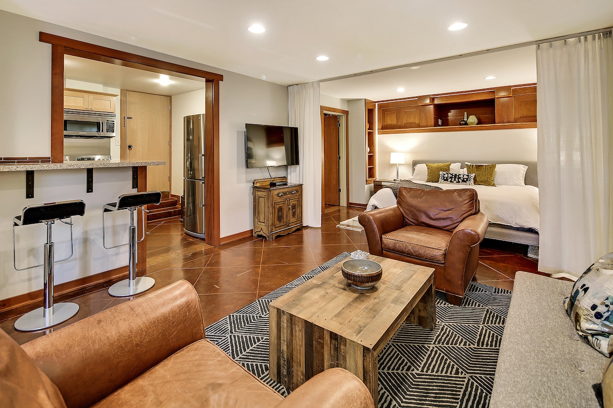 The open-plan living space presents a cozy seating area with two leather chairs and a rustic coffee table. A kitchen bar with two stools is visible, alongside a comfortable bed framed by built-in shelves. Soft lighting enhances the overall inviting atmosphere.