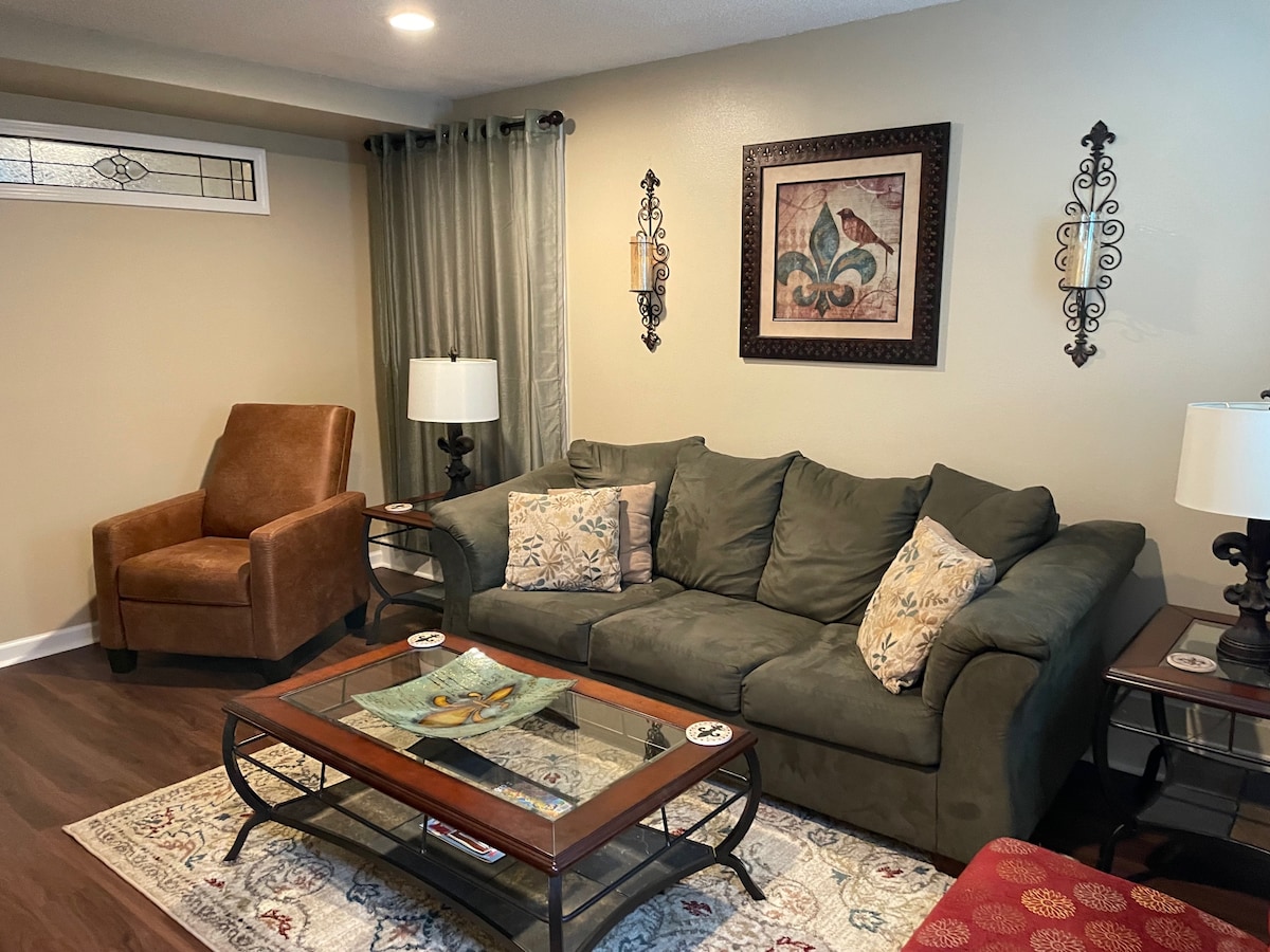 A warm living area features a comfortable green sofa adorned with decorative pillows. A wooden coffee table sits atop a patterned rug, displaying artwork. Two lamps provide soft lighting, while a brown armchair adds a touch of contrast against the light-colored walls.