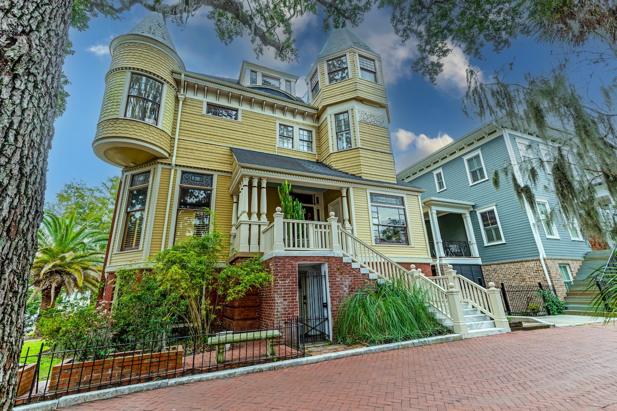 This historic Queen Anne Victorian mansion features a distinctive turret and unique architectural details. A welcoming front porch with decorative railings is approached by a brick pathway. Lush greenery and mature trees surround the home, highlighting its charm in a picturesque setting.