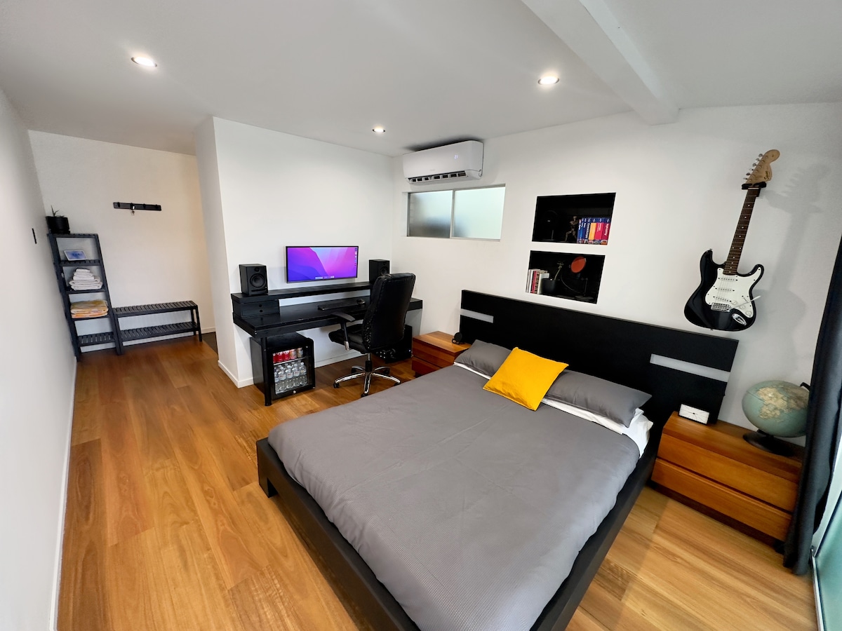 A modern guest suite features a spacious bed with a grey coverlet. A desk with a computer setup is positioned against the wall. Storage shelves display books and decorative items, while additional storage is available in a nearby unit. Natural light enters through a window.