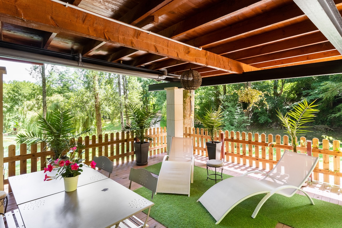 An outdoor terrace area features a set of lounge chairs and a dining table surrounded by greenery. Potted plants add a touch of nature, while a wooden fence provides privacy. A soft green rug enhances the inviting atmosphere.