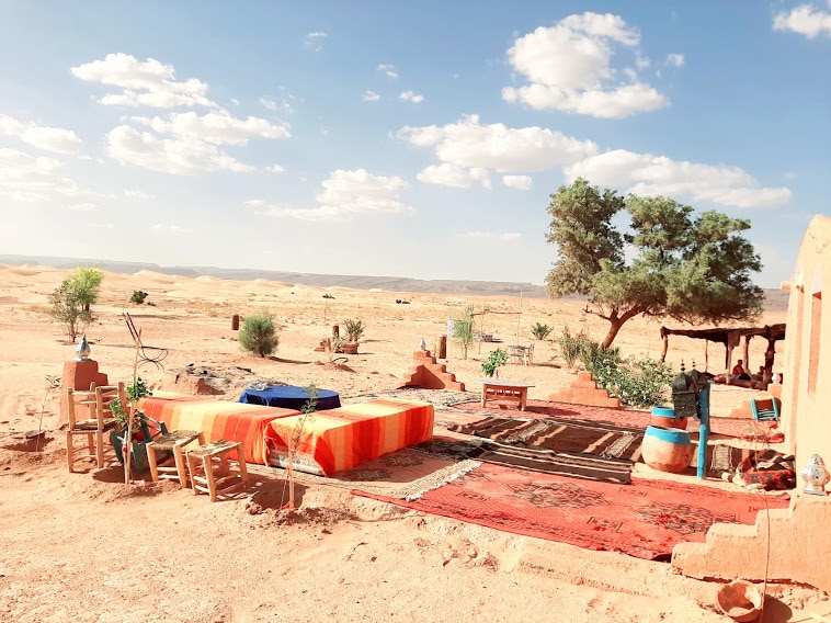 A serene outdoor area is presented, featuring an orange and white striped bed spread on a raised platform. Surrounding elements include wooden chairs, potted plants, and lush greenery, with gentle dunes visible in the background under a blue sky adorned with white clouds.