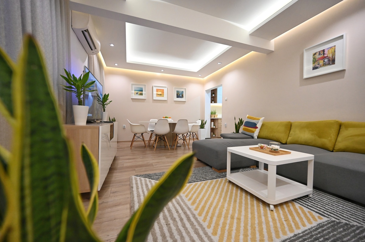 A spacious living room is furnished with a comfortable grey sofa and a coffee table. A bright dining area for six is visible in the background, complemented by indoor plants. Warm lighting highlights the neutral decor and enhances the inviting ambiance.