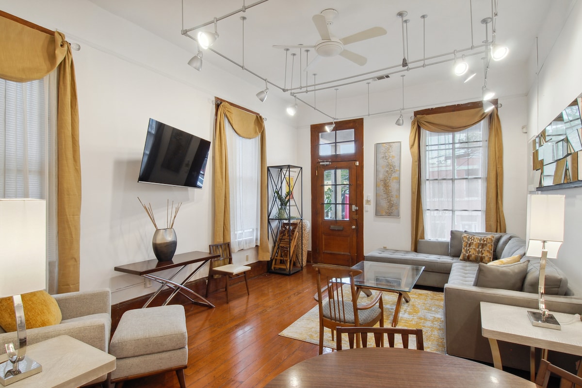 A bright living room is illuminated by natural light filtered through the large windows adorned with light-colored drapes. A comfortable sectional sofa and stylish coffee table are featured, along with a wall-mounted television. A mix of seating options, including armchairs, is present, enhancing the inviting space.