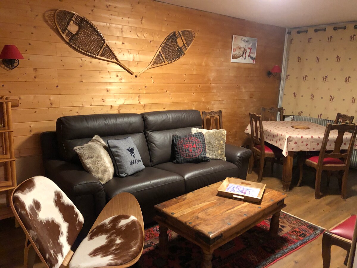 A warm living area features a dark leather couch adorned with decorative pillows. A rustic wooden coffee table sits atop colorful rugs, complemented by cowhide chairs. Wooden walls create a cozy atmosphere, with vintage snowshoes displayed above the couch.