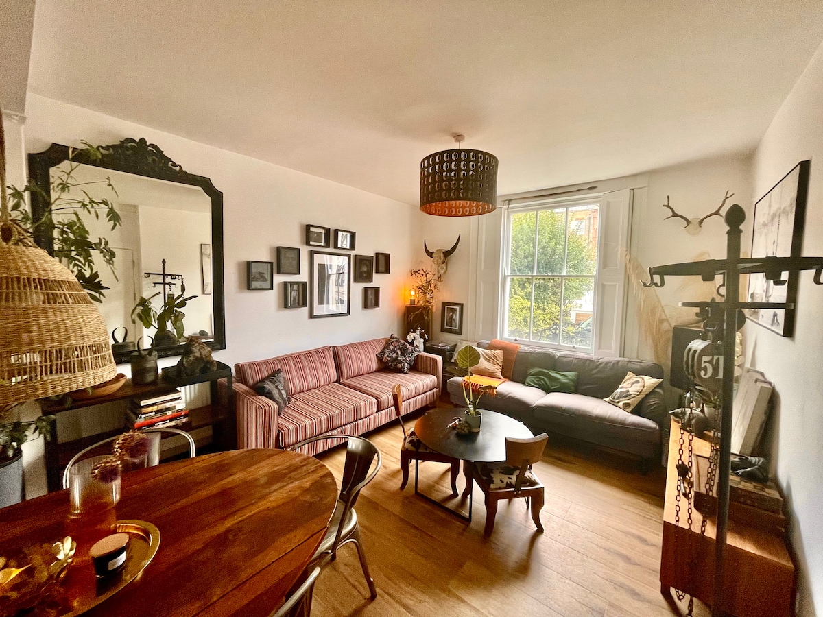 A welcoming living room is featured, with two sofas and a round table positioned in the center. The space is enriched by framed artwork on the walls and greenery displayed in various spots. Natural light illuminates through a large window, complementing the room's cozy ambiance.