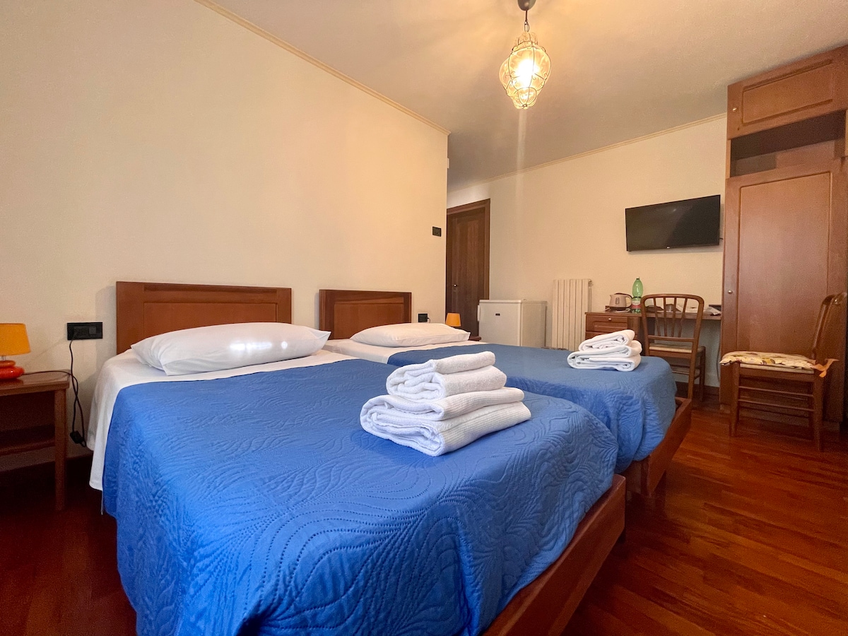 A spacious guest room features two single beds dressed in blue coverings. Neatly arranged white towels are placed at the foot of each bed. A desk and a flat-screen TV are visible, with warm wooden accents complementing the parquet flooring.