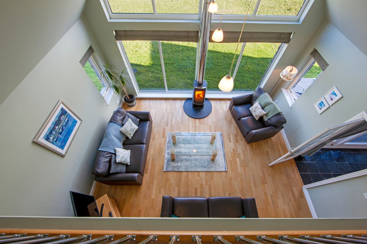 An airy lounge area is captured from above, showcasing comfortable seating arrangements around a glass coffee table. A log-burning stove is positioned centrally, with light streaming through large windows that frame the outdoor greenery. An inviting atmosphere is complemented by subtle decor elements.