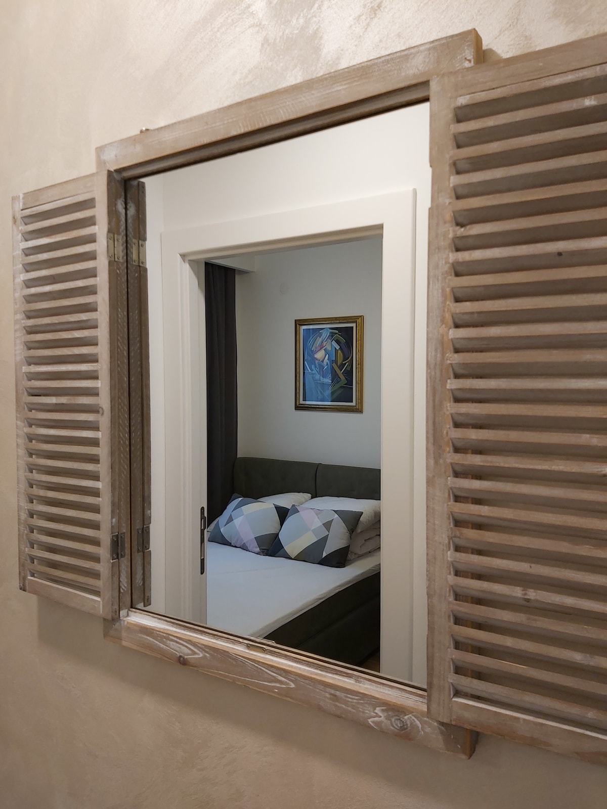 A framed mirror with wooden shutters reflects a cozy bedroom featuring a bed with geometric-patterned pillows and artwork hanging on the wall. The soft lighting enhances the inviting feel of the space.