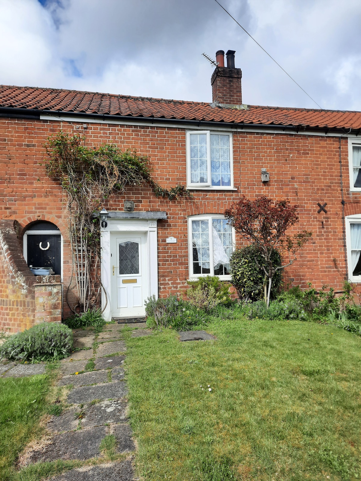 The exterior of a charming cottage is visible, featuring a red brick facade and white-framed windows. Lush greenery and colorful shrubs frame the entrance, complemented by a small path leading to the door. A traditional chimney rises above the roof under a partly cloudy sky.