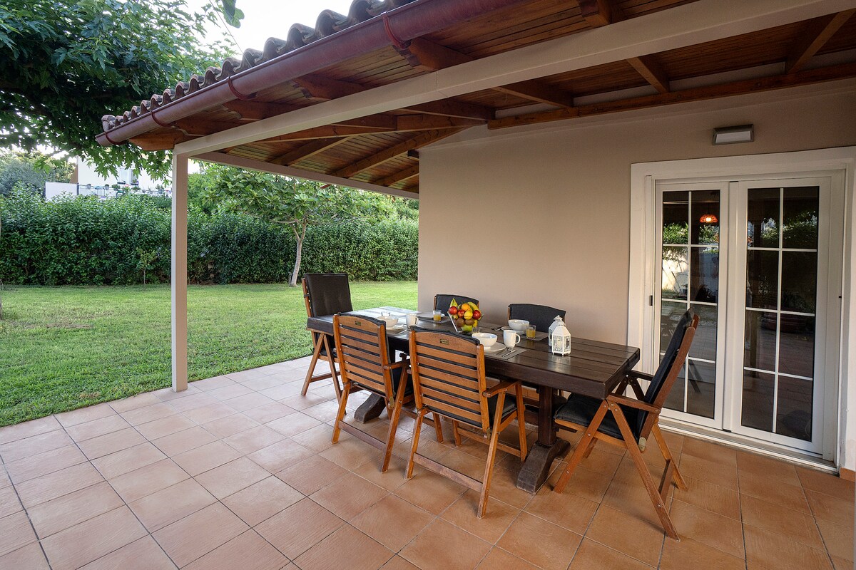 An outdoor terrace area features a wooden dining table surrounded by six chairs, set under a spacious roof. A garden is visible in the background, with lush green grass and trees creating a natural privacy barrier. Soft light illuminates the setting.