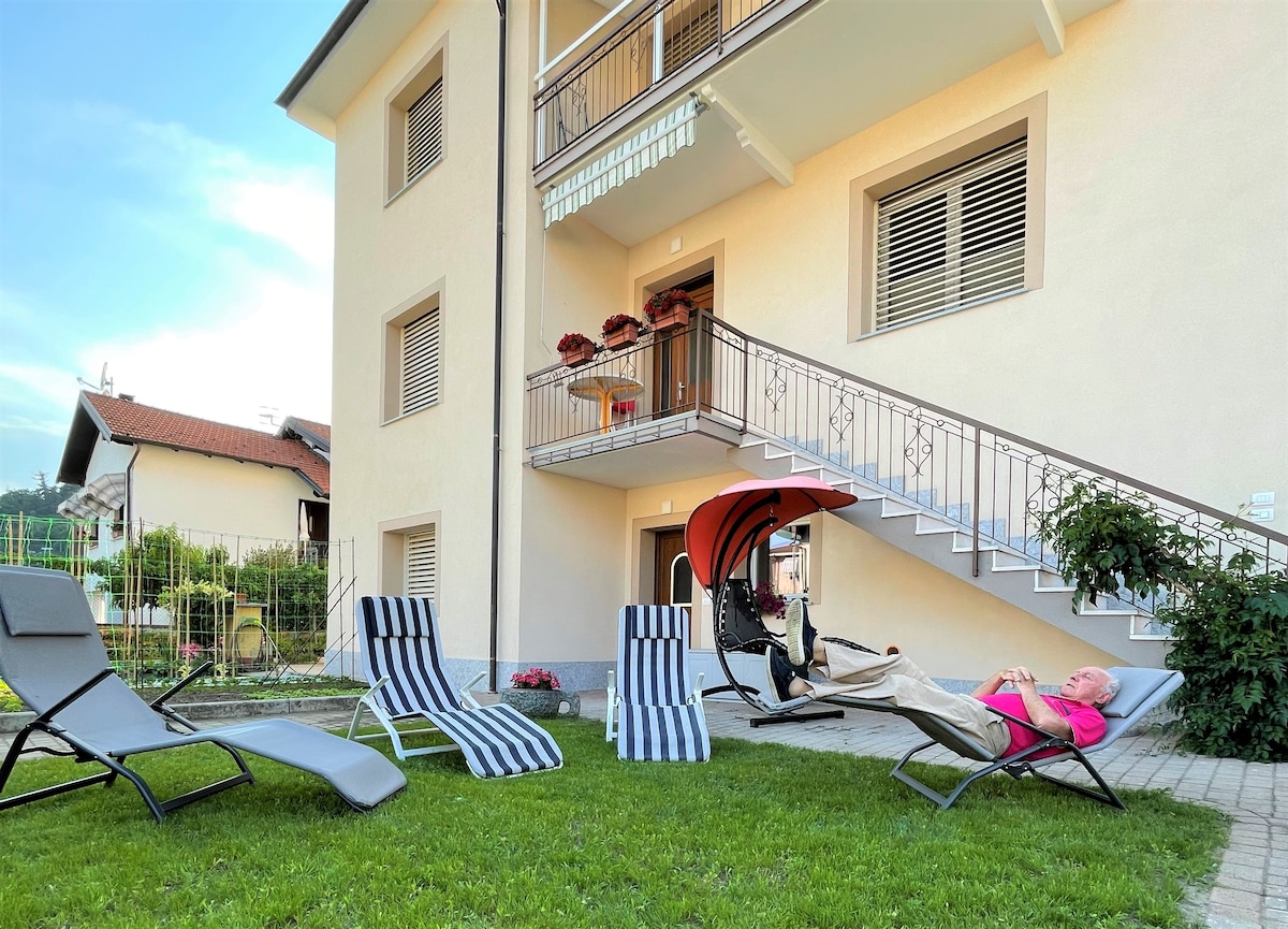 A spacious outdoor area is equipped with lounge chairs on a manicured lawn. A covered seating area is situated near the entrance of the building, providing shade. Flower pots adorn the balcony above, contributing to a pleasant, inviting environment.