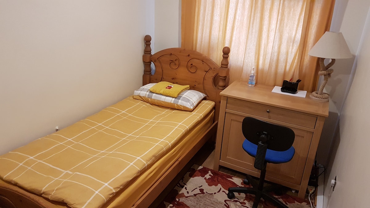 A single bed with a wood-framed headboard is positioned against the wall, topped with a yellow and white checkered blanket. A small desk with a blue chair is situated beside the bed, accompanied by a lamp and a bottle of water on the surface. Soft curtains filter natural light from a window.