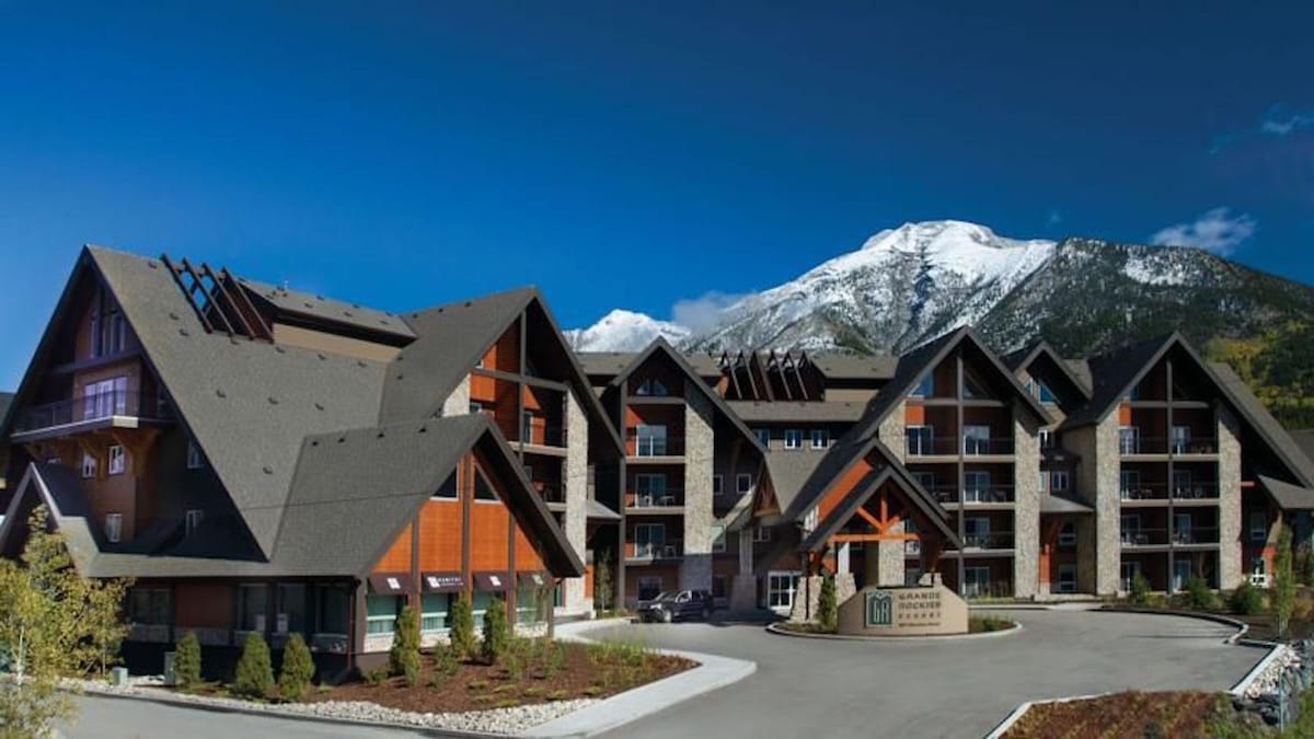 The exterior of the Grande Rockies Resort is showcased, featuring a combination of modern and rustic architectural styles. The structure is surrounded by landscaped gardens and a winding driveway, set against a backdrop of snow-capped mountains and a clear blue sky.