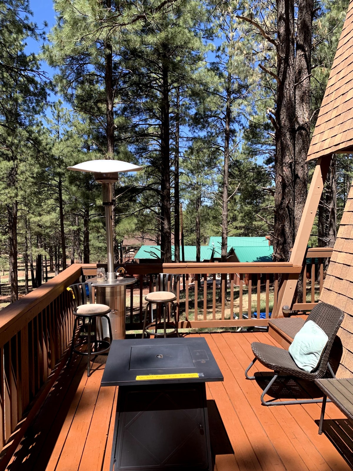 A wooden deck offers a tranquil outdoor space surrounded by tall pine trees. Two bar stools are positioned near a patio heater, and a small table sits adjacent to a lounge chair, creating a welcoming area for relaxation.