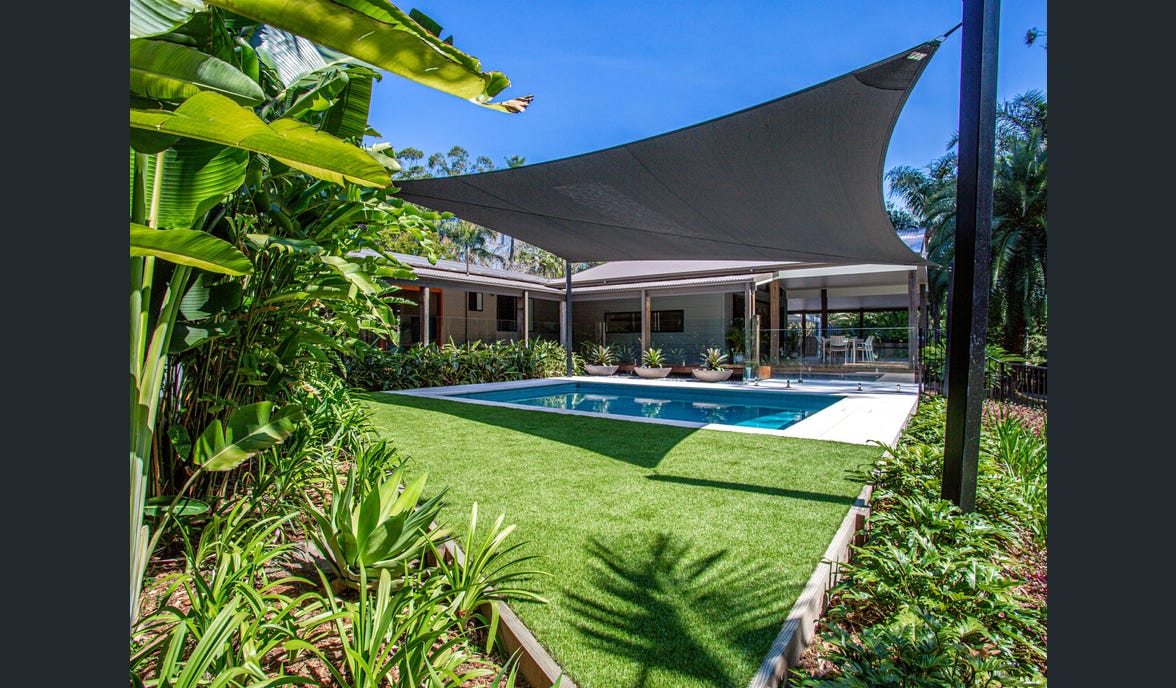 A shaded outdoor area surrounds a swimming pool, with vibrant green grass and tropical plants framing the space. The sleek pool reflects a clear blue sky, while the shade sail offers comfort from the sun, creating an inviting place for relaxation.