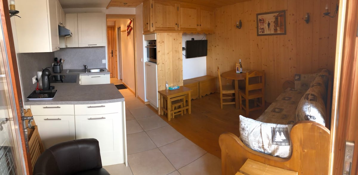 A modern kitchen area is situated next to a cozy living space featuring light wooden paneling. A dining table with four wooden chairs is positioned next to a comfortable sofa. A wall-mounted television is visible, and natural light enters through a window.