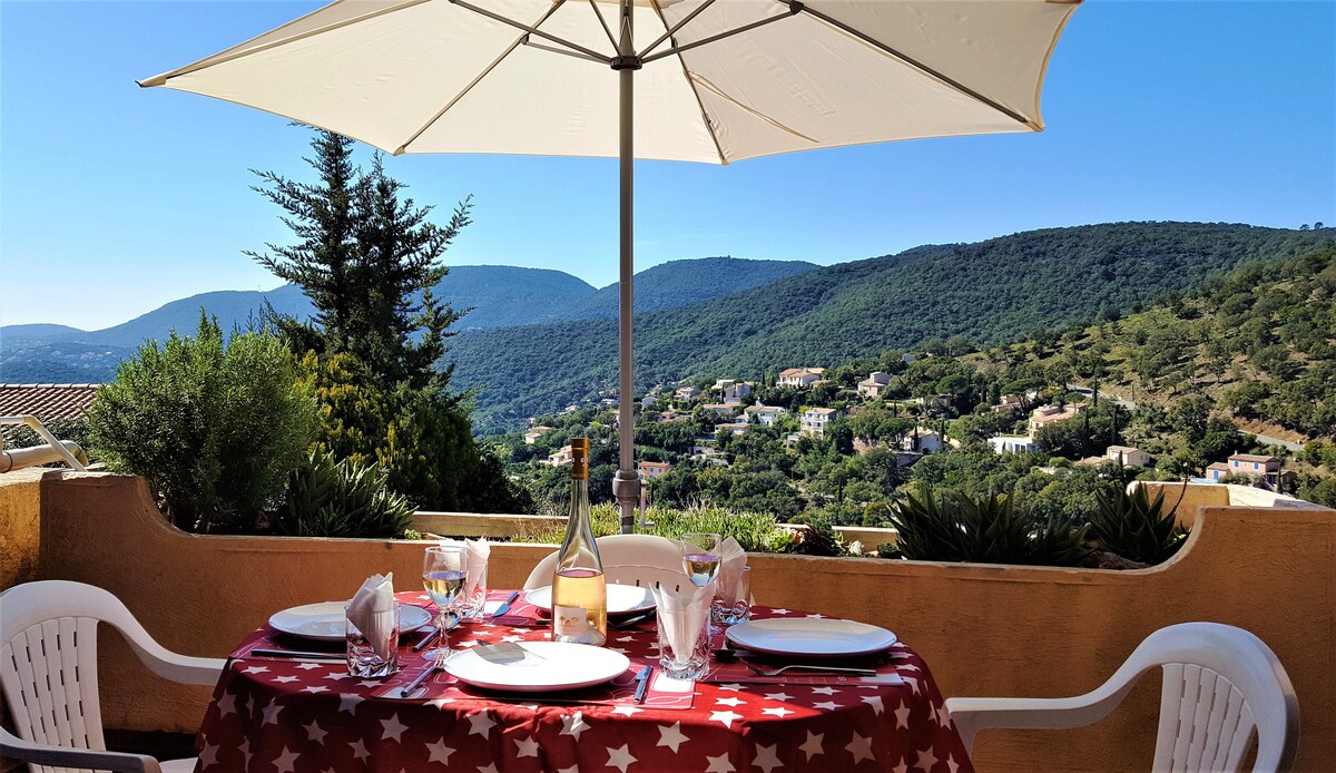 A terrace dining area is presented, featuring a round table covered with a star-patterned tablecloth. White chairs surround the table, with table settings and a bottle of wine placed in the center. The backdrop showcases lush green hills and clear blue skies.