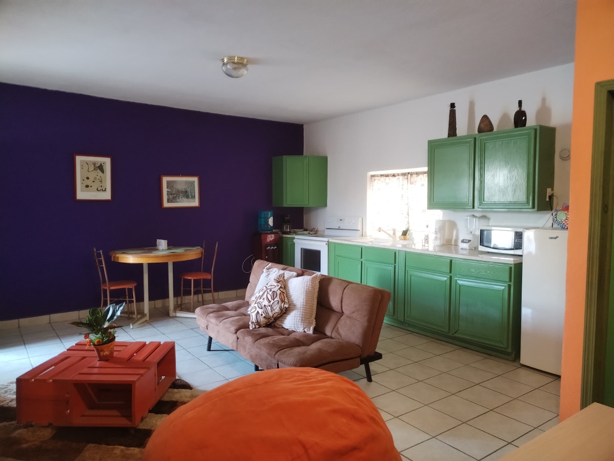 A spacious living area is presented, featuring a brown sofa bed and an orange bean bag. A small dining table is positioned near the window, while vibrant green kitchen cabinets are visible in the background. The purple wall adds an accent to the room.