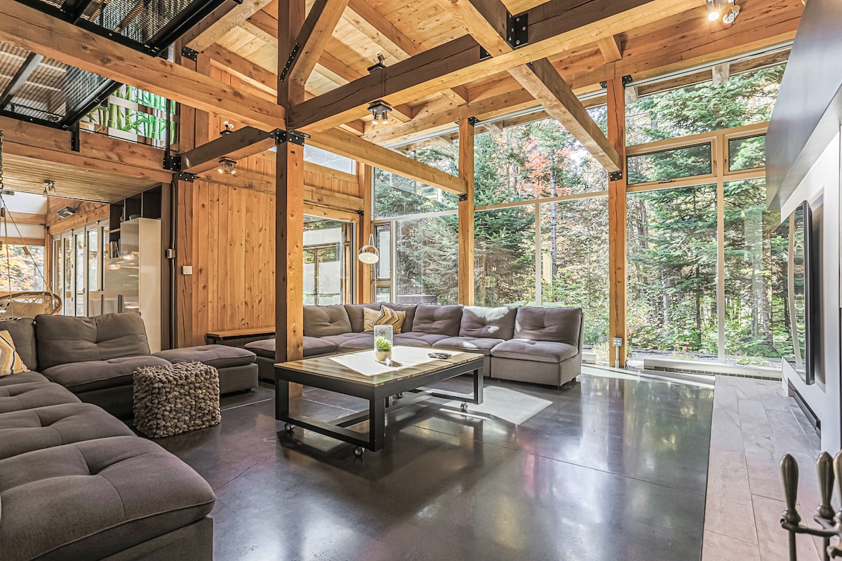 A spacious living area features wood beams and expansive windows, allowing natural light to illuminate the space. A large gray sectional sofa surrounds a central table, complemented by a cozy pouf. The view showcases a serene wooded area, enhancing the connection with nature.