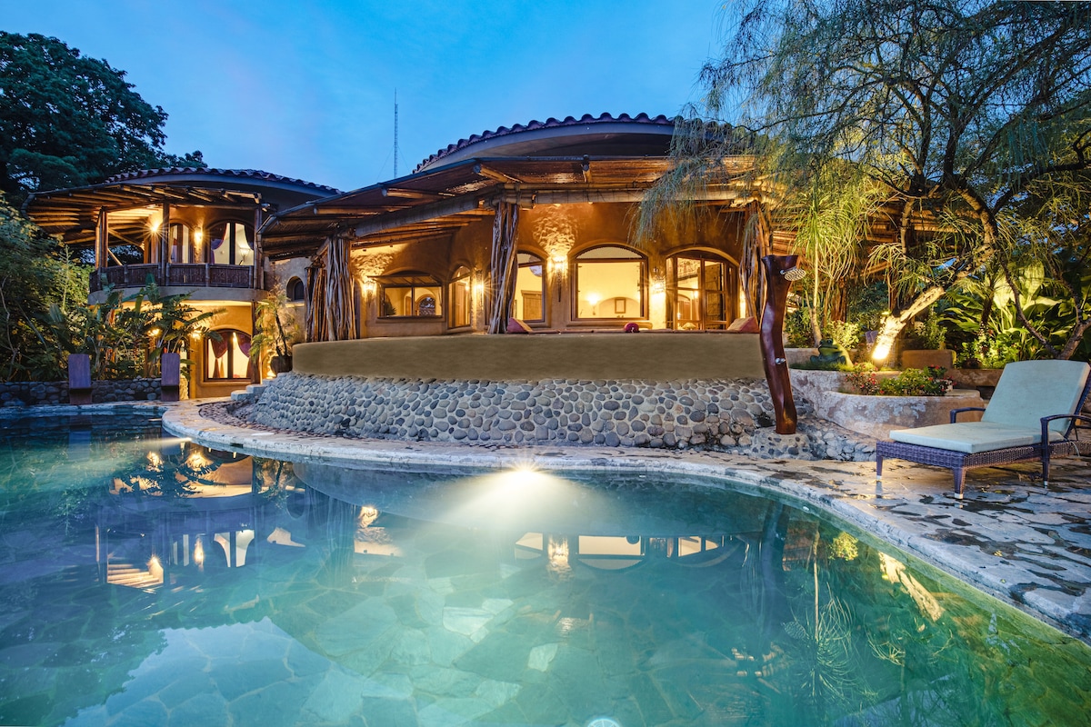 The villa's unique architectural design is complemented by lush greenery, featuring rounded stone shapes and wooden accents. An inviting infinity pool reflects the evening sky, while warm lights illuminate the exterior, creating a tranquil ambiance around the outdoor living space.