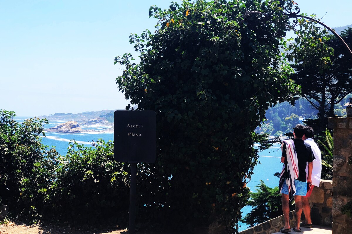 Two individuals are seen near a sign that reads 'Access Playa.' They stand beside lush greenery, and a scenic coastal view encompassing sandy shores and tranquil waters is visible in the background. The landscape features rolling hills and distant rocky formations, framed by foliage.