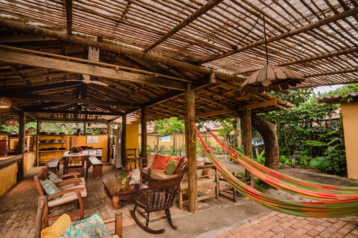 An open-air lounge area is highlighted by wooden beams and natural materials. Colorful cushions adorn the seating, while a hammock is suspended nearby. A spacious kitchen counter is partially visible in the background, surrounded by lush greenery and warm-toned walls.