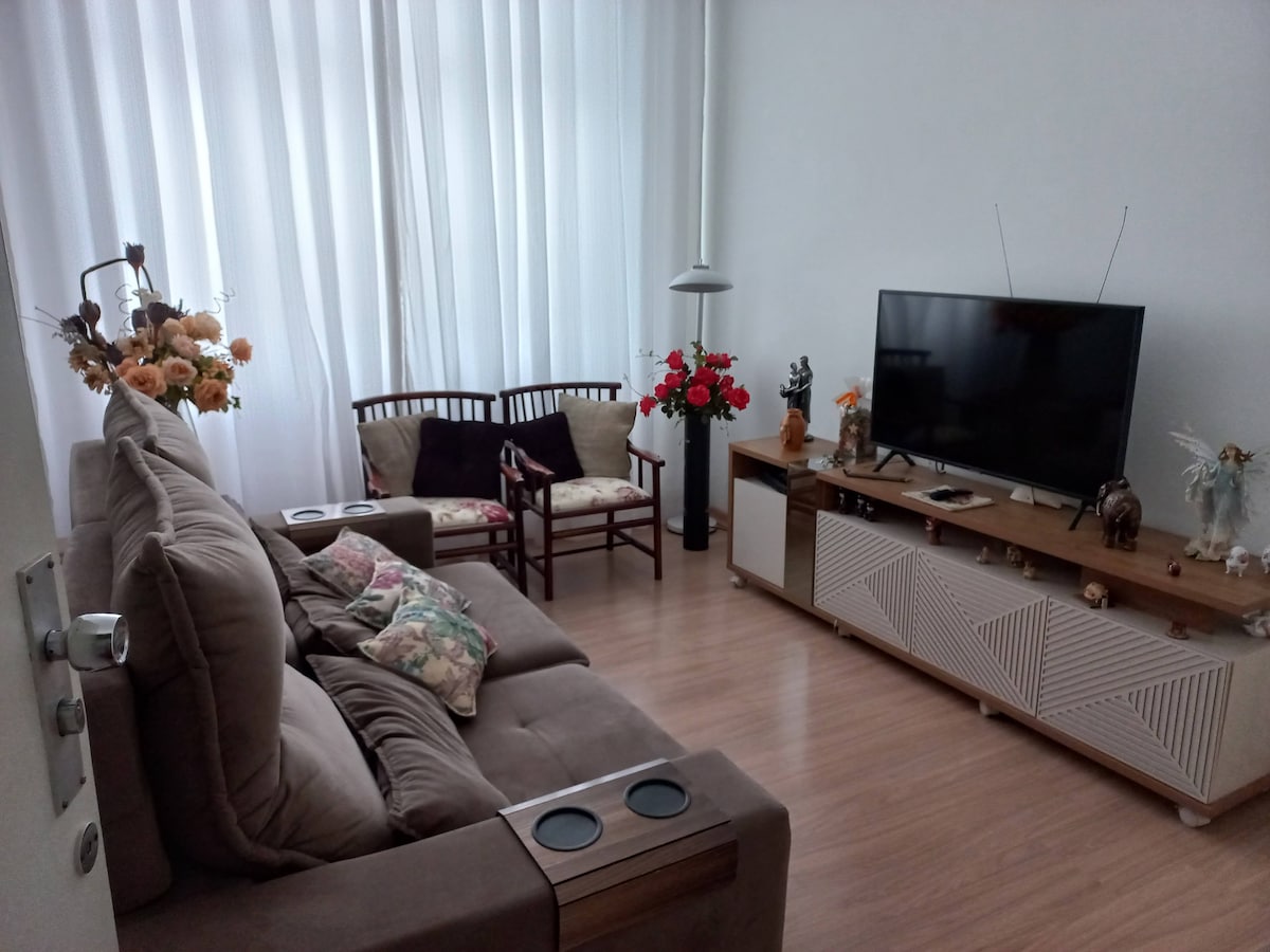 The living area features a comfortable large sofa with built-in cup holders and decorative pillows. A wooden TV stand supports a flat-screen television, while a small wooden chair set is positioned nearby. Natural light filters through sheer white curtains, and flowers add a touch of color.