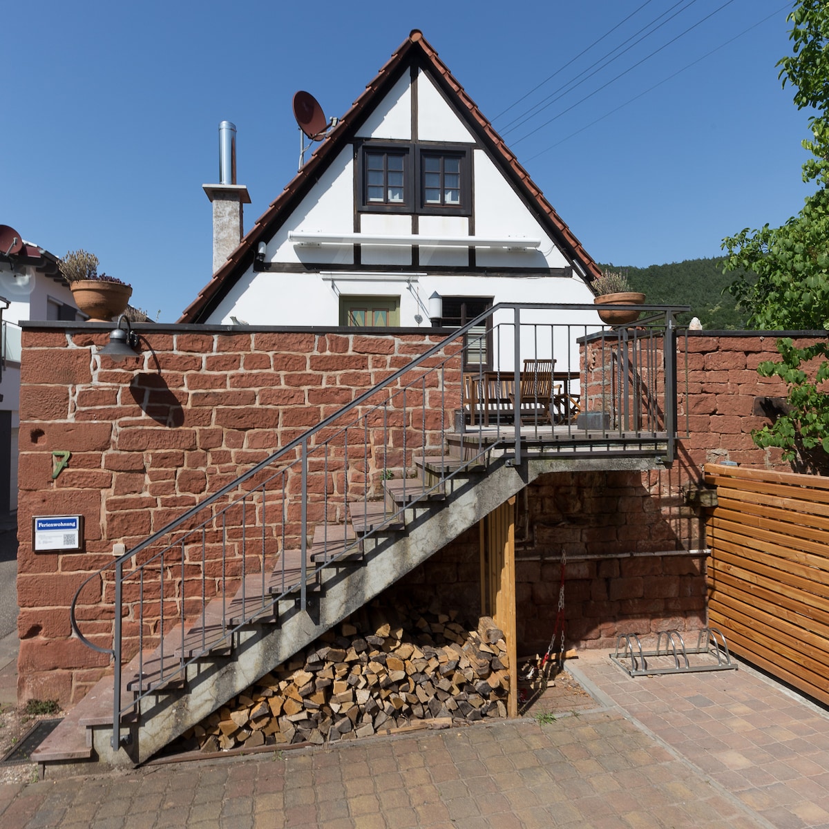 An exterior view of a two-story house showcases a staircase leading to a balcony. The entrance is surrounded by stone walls, and outdoor seating is visible on the balcony. Firewood is stacked neatly at the base of the stairs, complemented by greenery.
