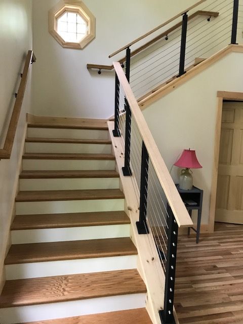 The staircase features natural wood steps and railings, enhanced by black metal accents. A small table with a pink lamp is positioned at the base, while a round window allows light to filter in. The surrounding walls are painted a soft hue.