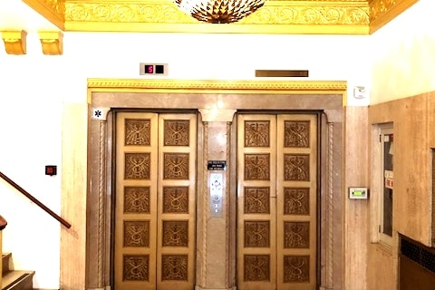 Two ornate elevators are framed by decorative golden trim and intricate detailing on the doors. Above, a chandelier adds elegance to the space, while illuminated floor indicators are visible beside each elevator. The surrounding walls are adorned with light-colored stone finishes.