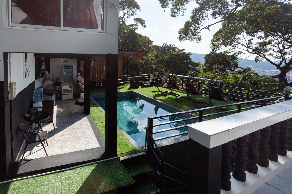A view of the outdoor area showcases a swimming pool surrounded by lush greenery and lounge chairs. The adjoining indoor space features a large window, complementing the inviting atmosphere. Sunlight filters through the trees, creating a serene environment.